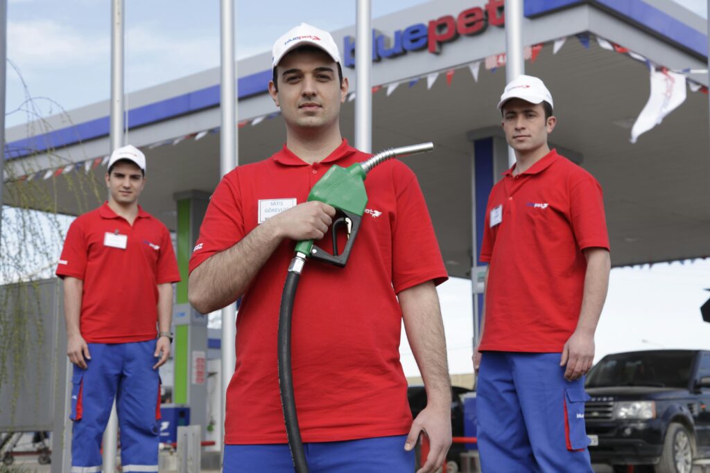 Three gas station attendants posing with a fuel nozzle at an outdoor petrol station.